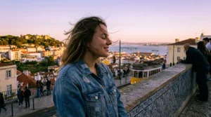 Eine junge Frau betrachtet den Sonnenuntergang über Lissabon von einem Miradouro aus, mit der Brücke des 25. April und dem Tejo im Hintergrund.